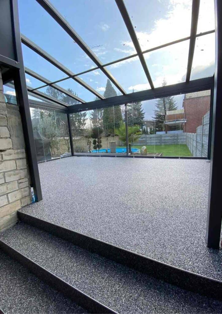 Moderner Steinteppich auf einem Balkon mit Glasüberdachung und Blick ins Gartengelände.